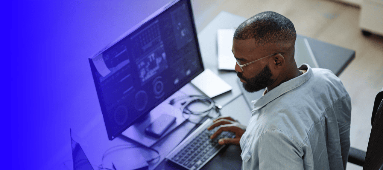 AI expert working at a desk, typing on a keyboard while viewing data dashboards on a widescreen monitor, with a blue gradient overlay on the left side of the image.