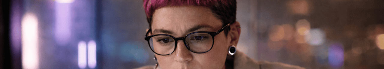 Close-up of a AI creative professional with short pink hair and glasses, focused on a screen in a softly lit workspace.