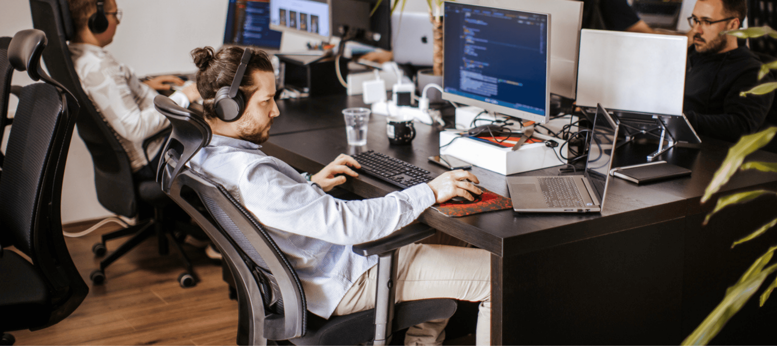A focused professional wearing headphones works across multiple monitors displaying code in a modern, open office, surrounded by laptops and technology that support a productive digital workspace.