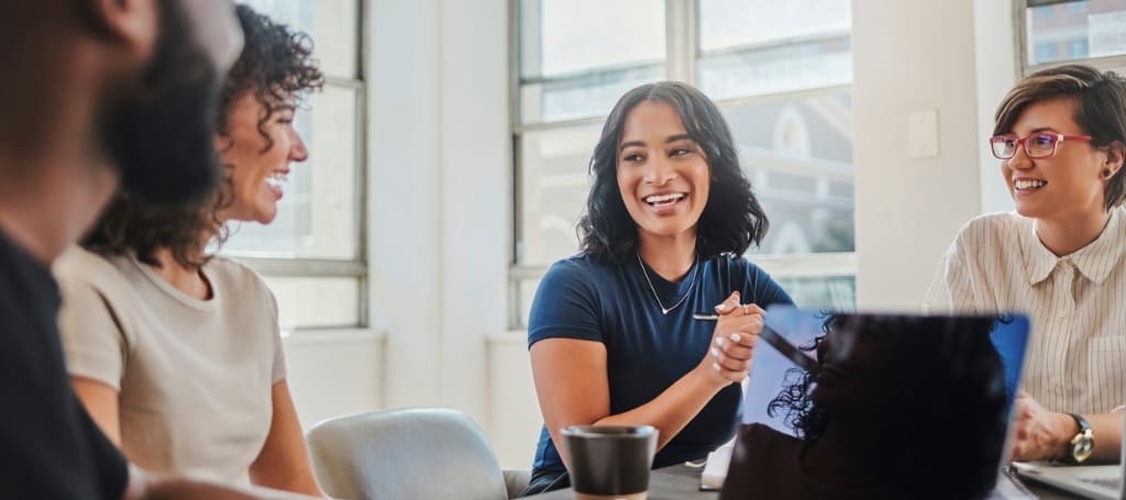 Business meeting with four young in-house design employees sitting at table smiling at each other and looking at laptop