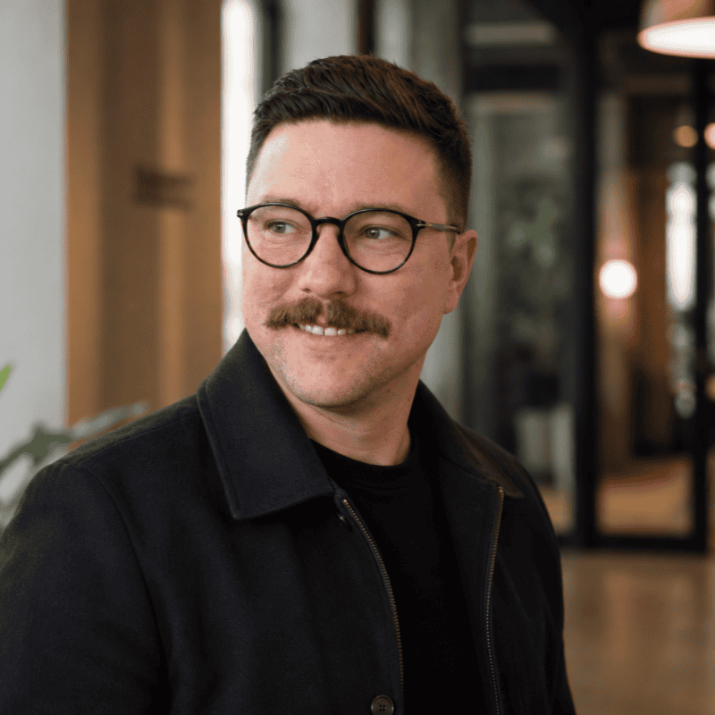 Headshot of Lowman Harley, Director of AI Enablement, a person with short dark hair, round glasses, and a mustache, smiling and looking to the side in a modern office setting.