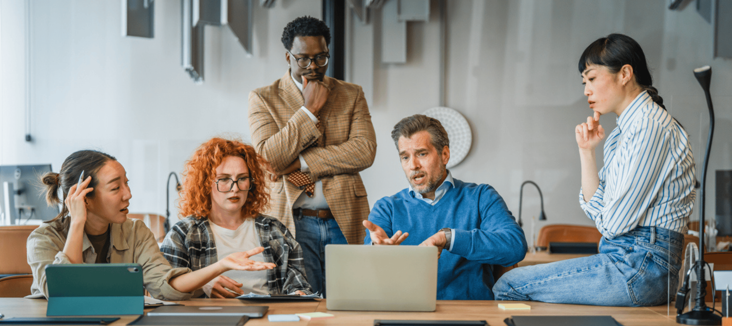 A diverse team of professionals gathers around a laptop in a collaborative office setting, discussing a complex project and exchanging ideas. Their focused expressions and active conversation convey teamwork, problem-solving, and strategic decision-making.